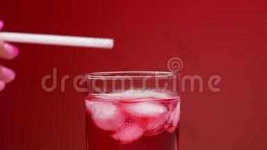 一个女人`她的手搅动冰块在一杯冰和红色苏打水与气体。 红色背景。 慢速饮料概念